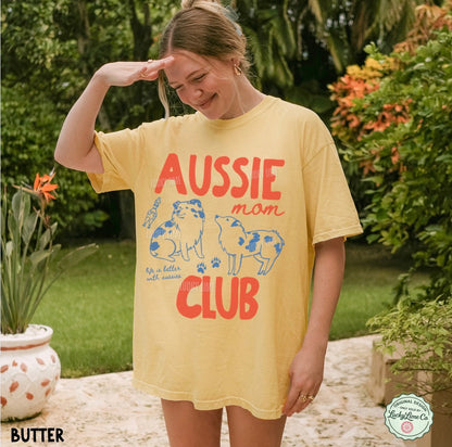 A woman wearing a yellow graphic t-shirt with 'AUSSIE mom CLUB' printed in red and blue, standing outdoors in a garden setting.