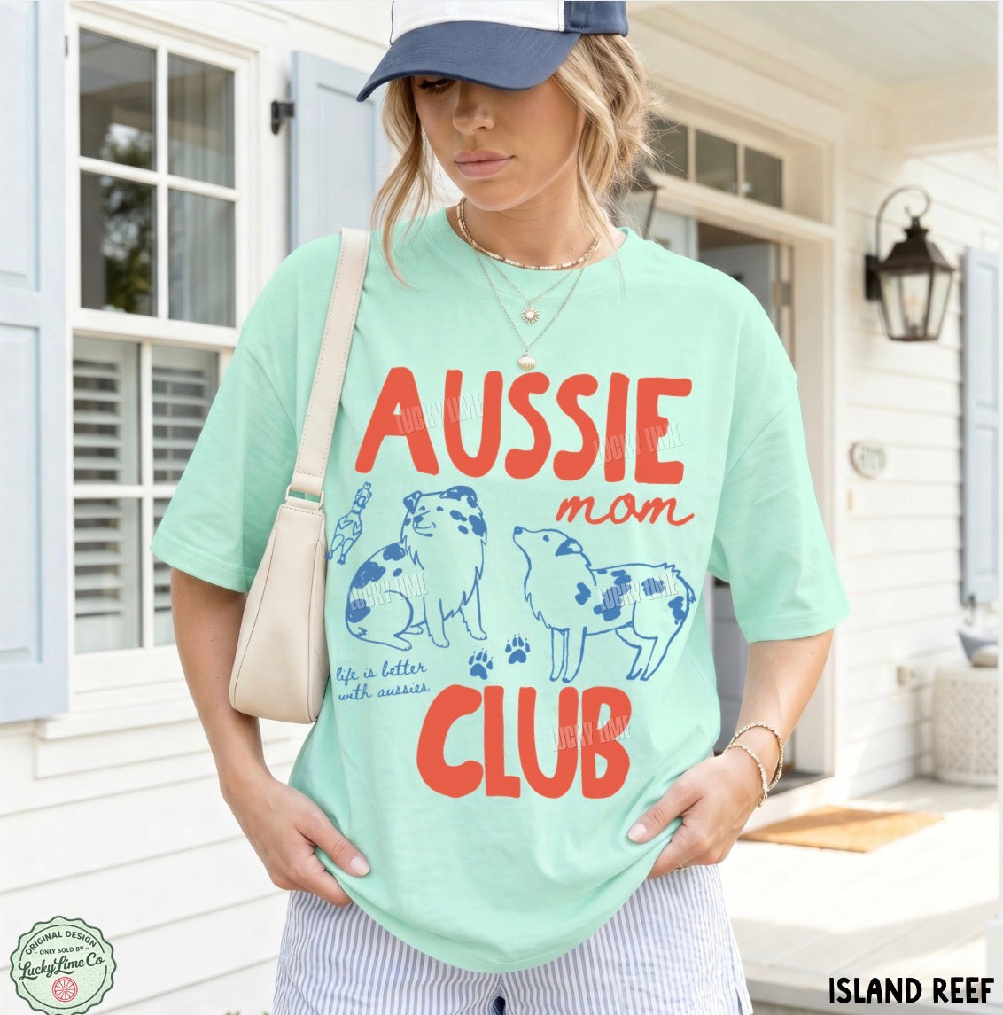 A woman wearing a mint green graphic t-shirt with 'AUSSIE mom CLUB' printed in red and blue, paired with a navy cap and a cream shoulder bag, standing in front of a white house porch.