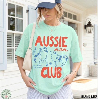 A woman wearing a mint green graphic t-shirt with 'AUSSIE mom CLUB' printed in red and blue, paired with a navy cap and a cream shoulder bag, standing in front of a white house porch.