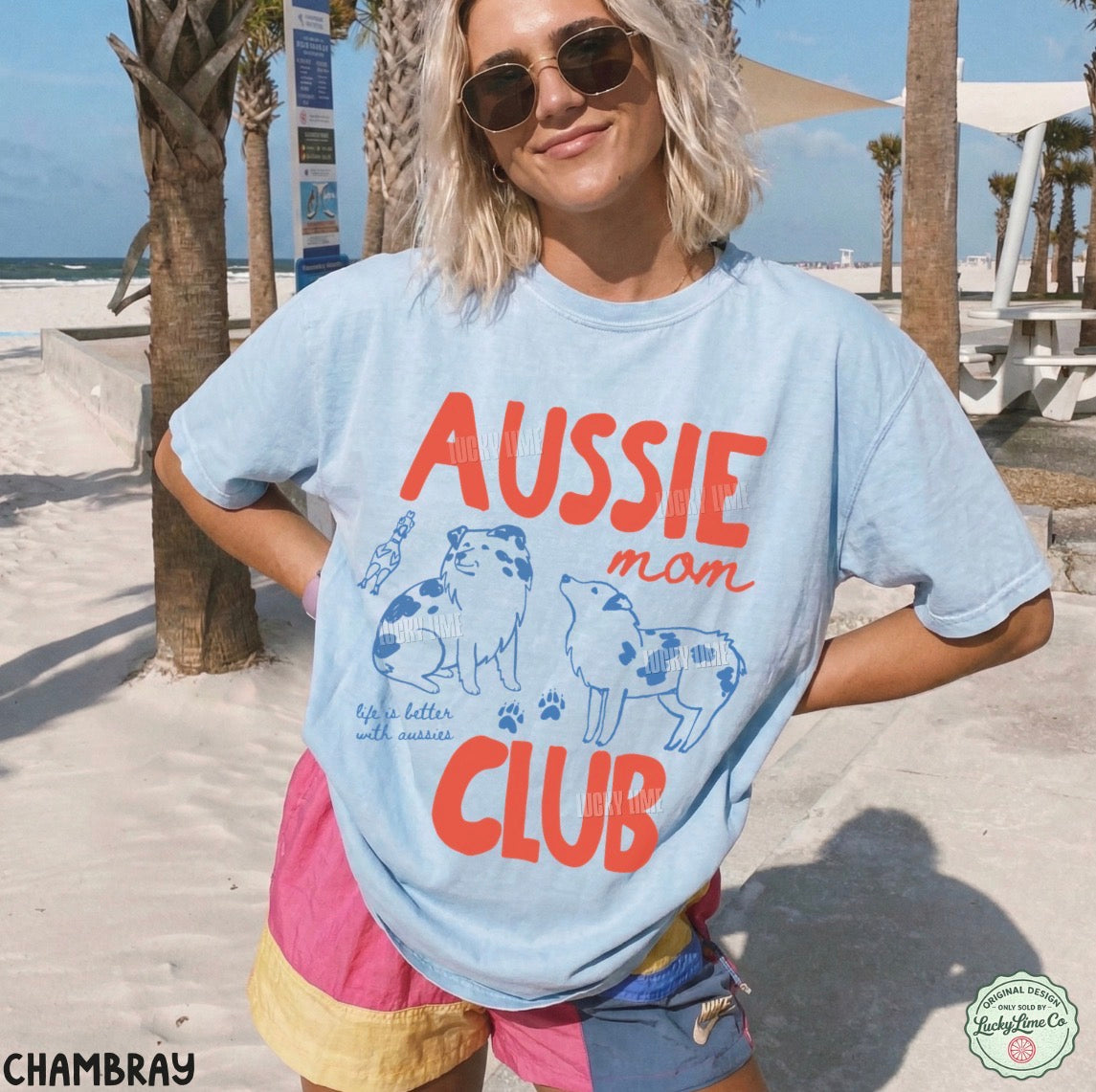 A woman wearing a light blue graphic t-shirt with 'AUSSIE mom CLUB' printed in orange and blue on the front, standing on a sunny beach with palm trees and sand in the background.