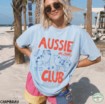 A woman wearing a light blue graphic t-shirt with 'AUSSIE mom CLUB' printed in orange and blue on the front, standing on a sunny beach with palm trees and sand in the background.