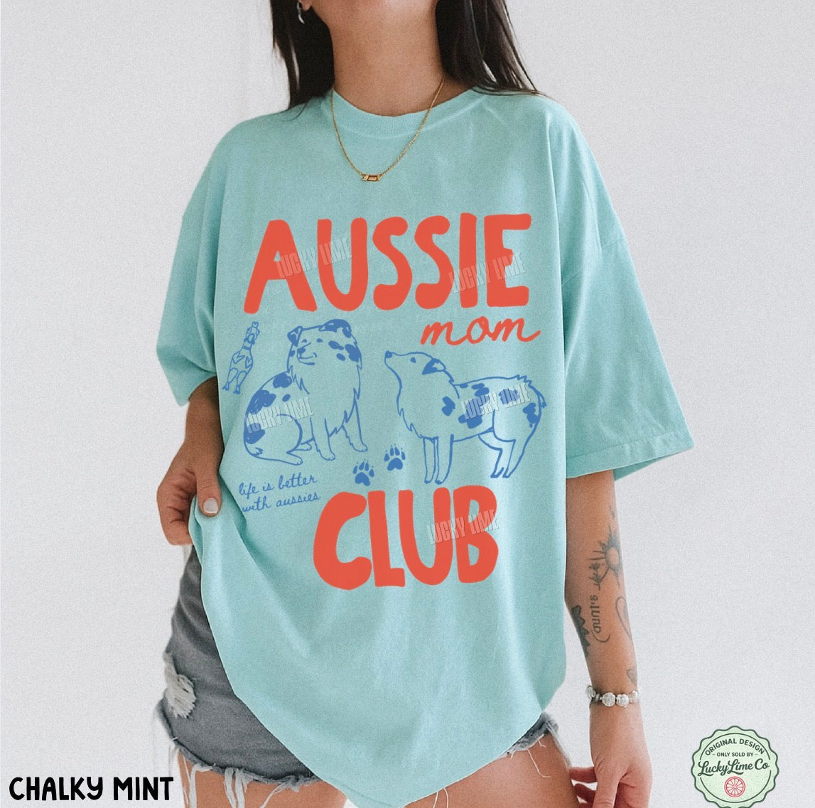A woman wearing an oversized light mint blue graphic T-shirt with orange and blue dog illustrations and the words 'AUSSIE mom CLUB'. The photo is cropped from the neck down against a plain light background, with 'CHALKY MINT' text at the bottom left and a circular logo at the bottom right.