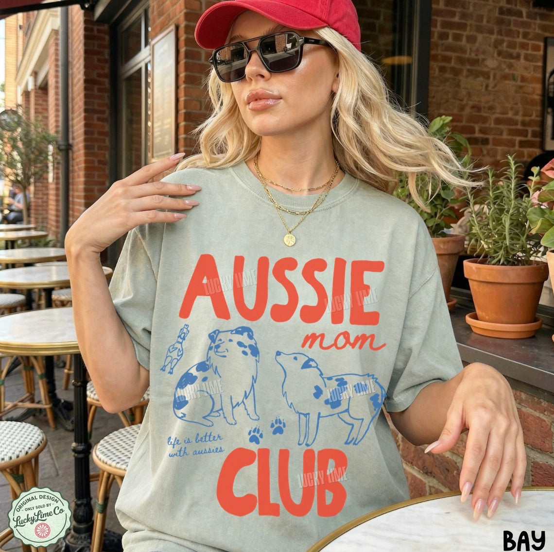 A woman wearing a light sage green graphic t-shirt with 'AUSSIE mom CLUB' printed in red and blue, paired with a red cap and sunglasses, posing at an outdoor cafe.