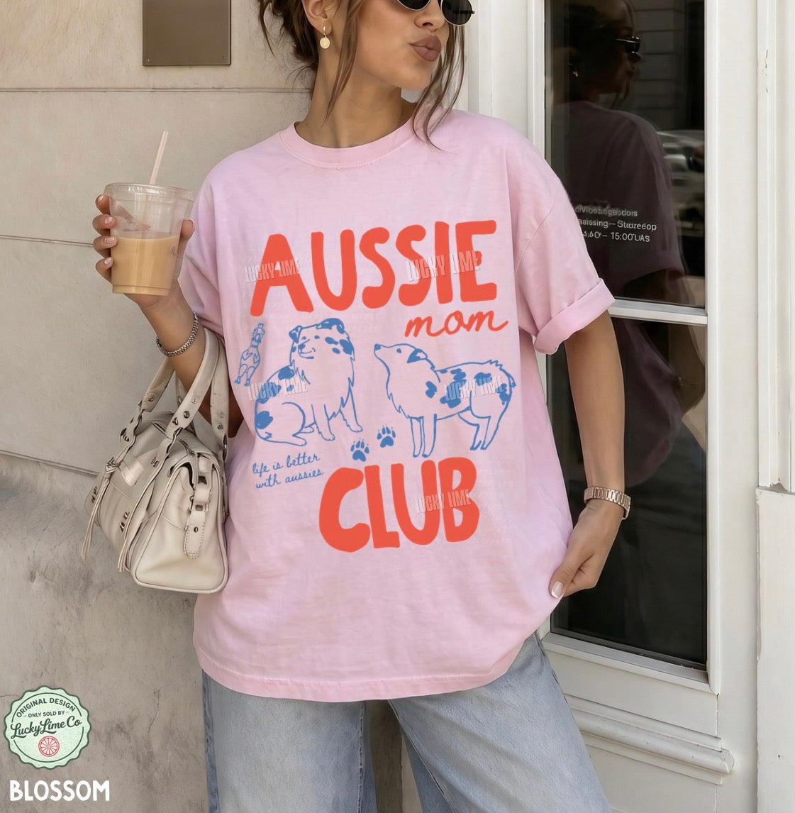 A woman wearing an oversized light pink graphic T-shirt with orange and blue text reading 'AUSSIE mom CLUB' and illustrations of two dogs, holding a drink and carrying a white handbag in an outdoor storefront setting.