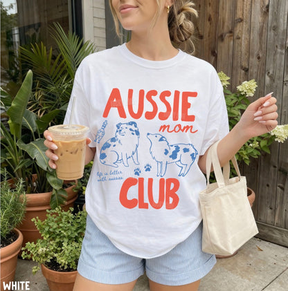 A woman wearing a white graphic t-shirt with 'AUSSIE mom CLUB' printed in orange and blue, holding an iced coffee and a canvas tote bag in front of a wooden fence and potted plants.