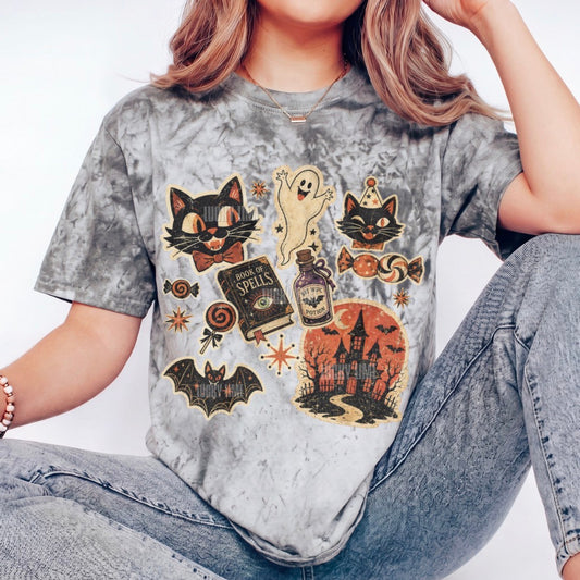 A person wearing a gray smoke tie-dye t-shirt with a Halloween graphic print featuring cats, a ghost, a potion bottle, and a haunted house, paired with light-wash jeans.