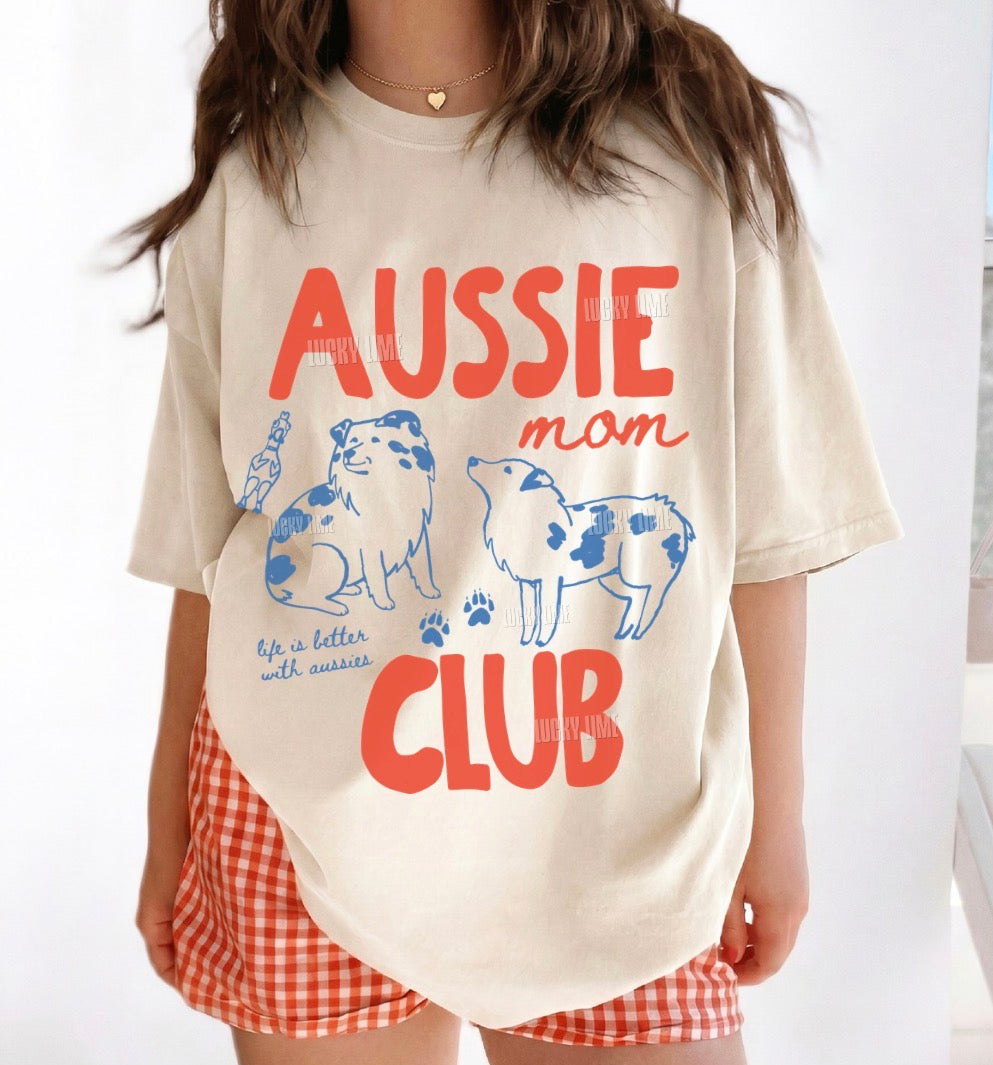 A woman wearing an oversized cream-colored t-shirt with a blue and red graphic that reads 'AUSSIE mom CLUB' with illustrations of two dogs. She is also wearing red and white gingham shorts.