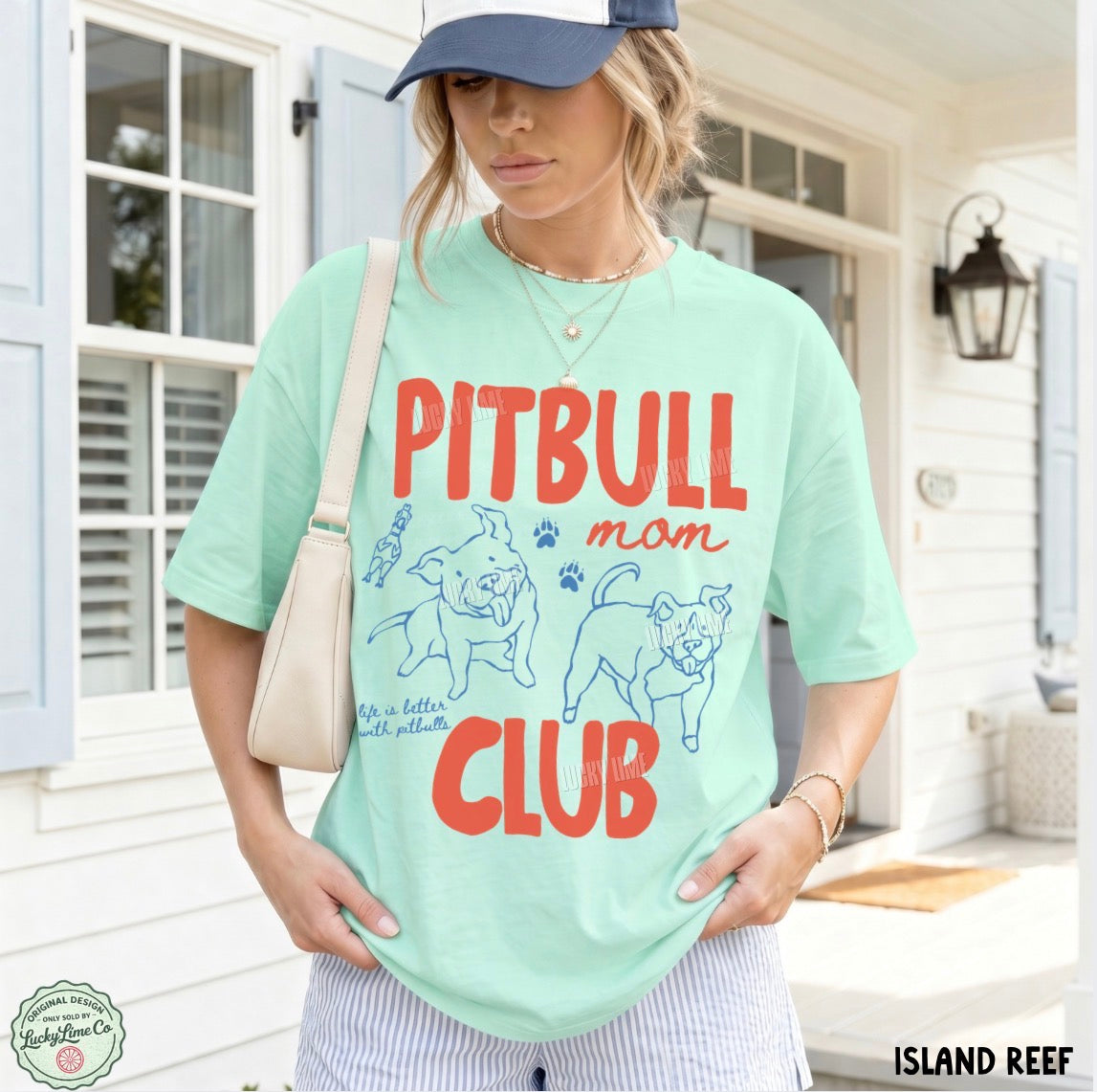 A woman wearing a mint green graphic t-shirt with 'PITBULL mom CLUB' printed in red and blue, paired with a navy blue baseball cap, a cream shoulder bag, and striped pants, standing in front of a white house porch.