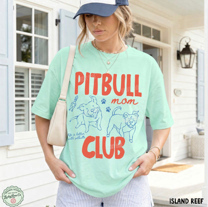 A woman wearing a mint green graphic t-shirt with 'PITBULL mom CLUB' printed in red and blue, paired with a navy blue baseball cap, a cream shoulder bag, and striped pants, standing in front of a white house porch.