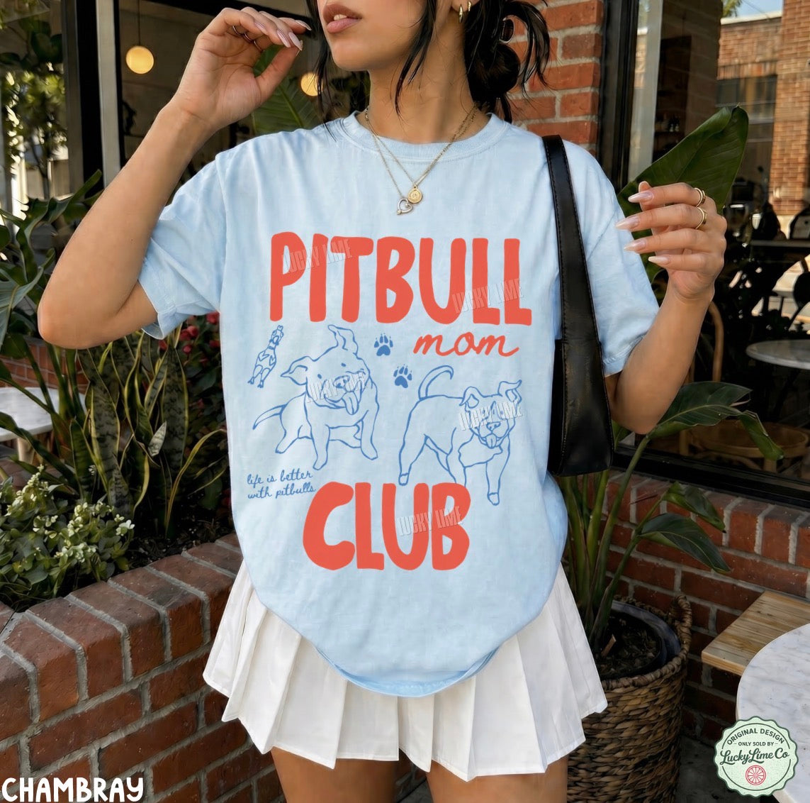 A woman wearing a light blue graphic T-shirt with 'PITBULL mom CLUB' printed in red and blue, paired with a white pleated skirt, photographed outdoors near a brick wall and plants.