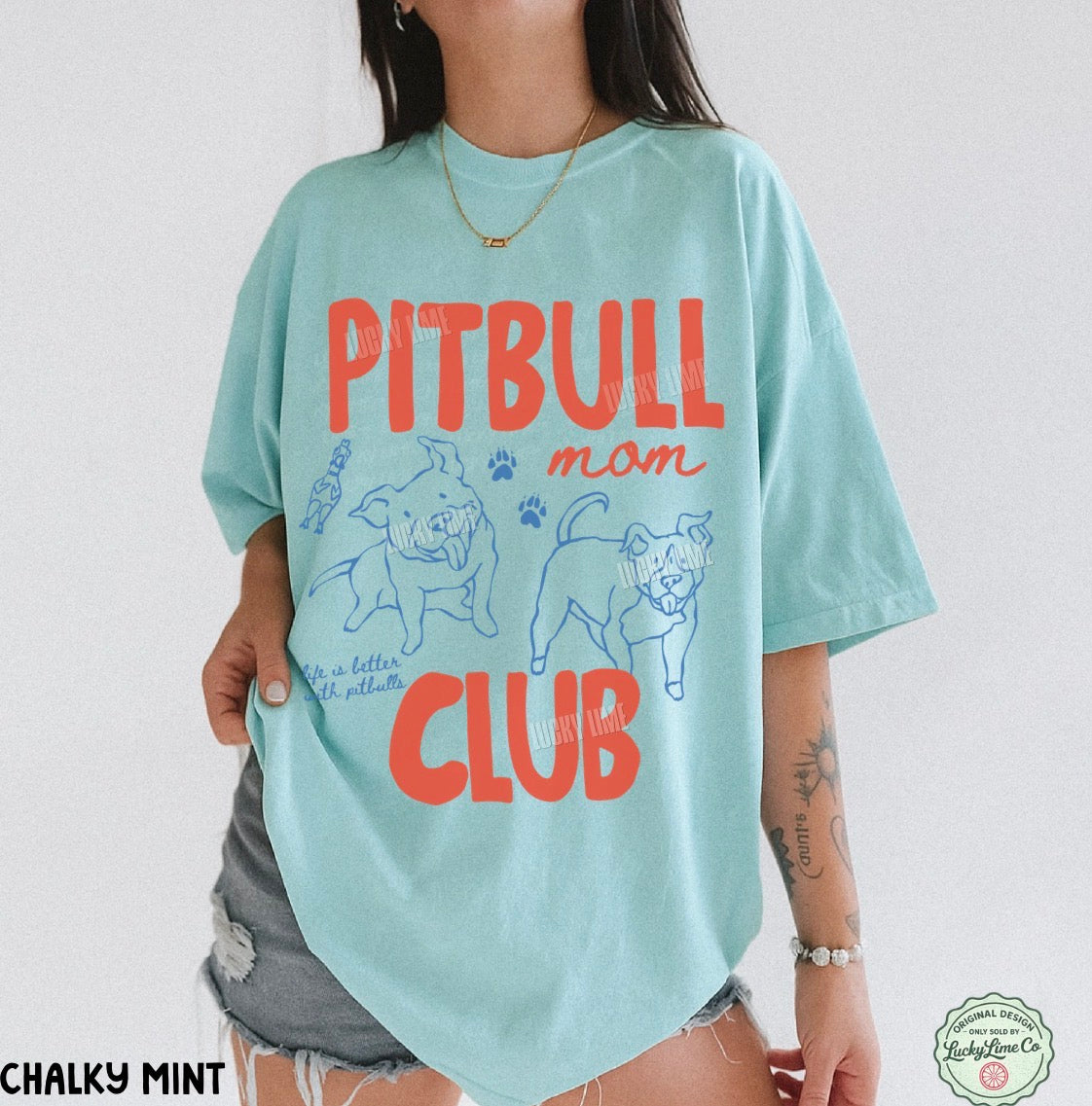 A woman wearing an oversized light mint green graphic t-shirt with 'PITBULL mom CLUB' printed in orange-red and blue on the front, paired with gray denim shorts.