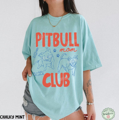 A woman wearing an oversized light mint green graphic t-shirt with 'PITBULL mom CLUB' printed in orange-red and blue on the front, paired with gray denim shorts.