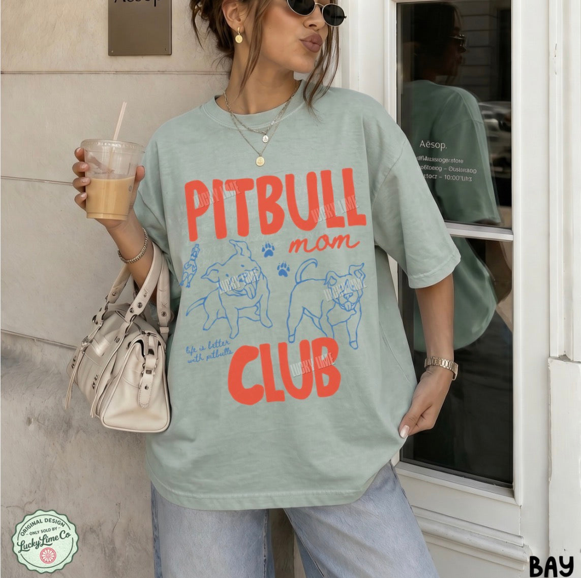 A woman wearing an oversized sage green graphic t-shirt with 'PITBULL mom CLUB' printed in red and blue, holding an iced coffee and a cream handbag.