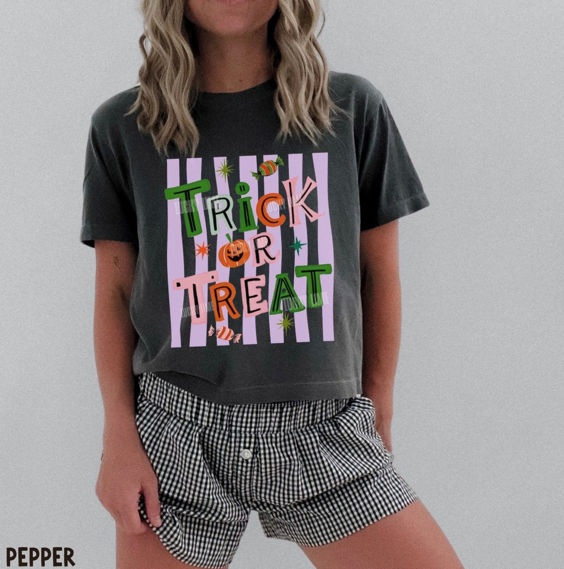 A woman wearing a dark charcoal gray graphic t-shirt with a 'Trick or Treat' Halloween design and black-and-white gingham shorts, standing against a light gray background.
