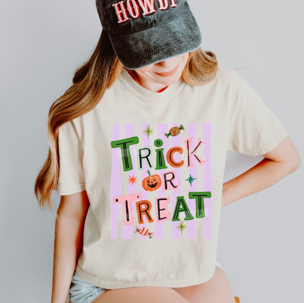 A woman wearing a cream-colored t-shirt with a colorful 'Trick or Treat' graphic print, paired with a dark gray cap that says 'HOWDY'.