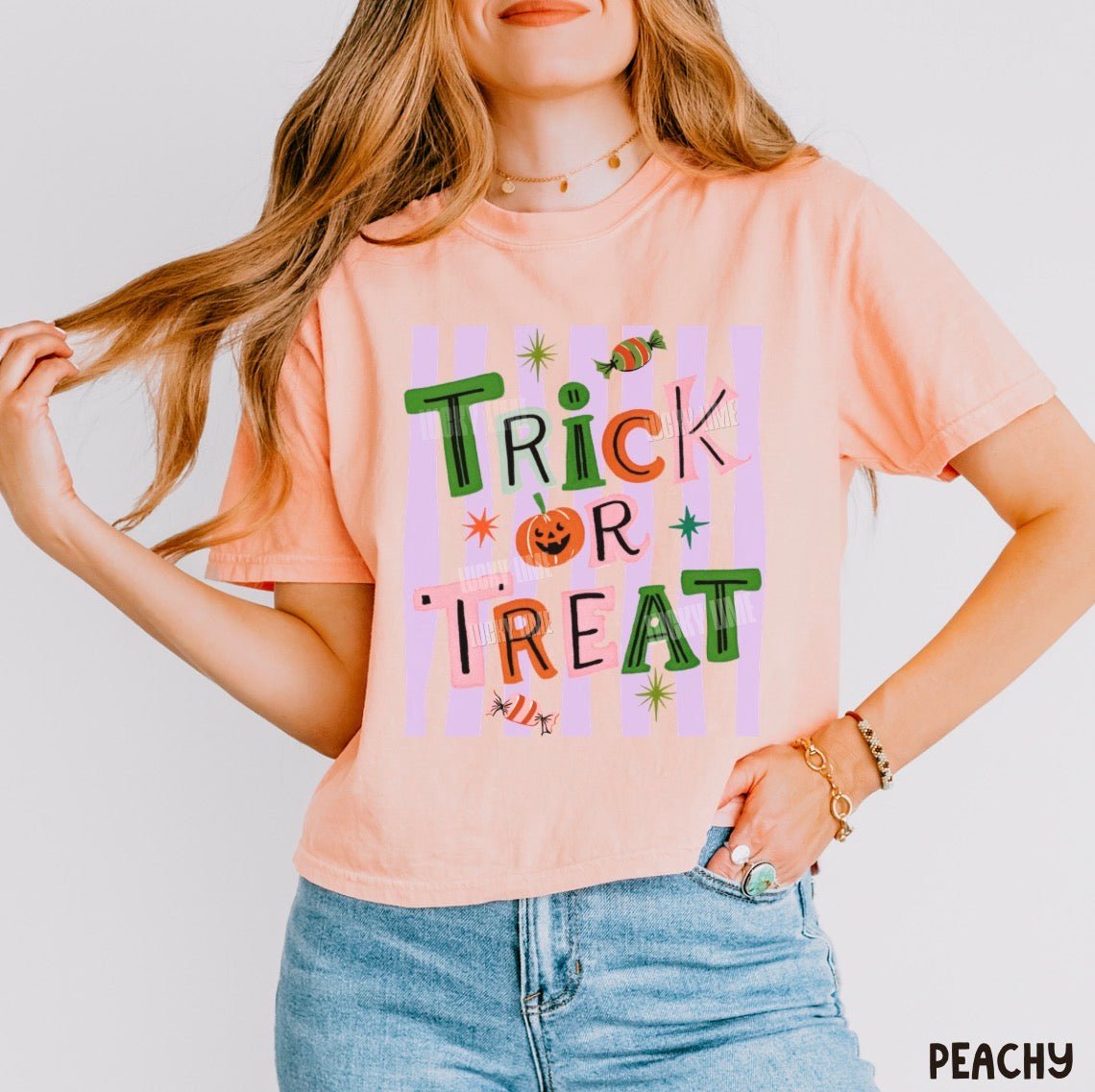 A woman wearing a peach-colored graphic t-shirt with 'Trick or Treat' written in colorful, playful letters over pastel pink and purple stripes. She is also wearing light blue denim jeans and gold jewelry, posing against a clean white background.