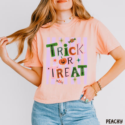 A woman wearing a peach-colored graphic t-shirt with 'Trick or Treat' written in colorful, playful letters over pastel pink and purple stripes. She is also wearing light blue denim jeans and gold jewelry, posing against a clean white background.