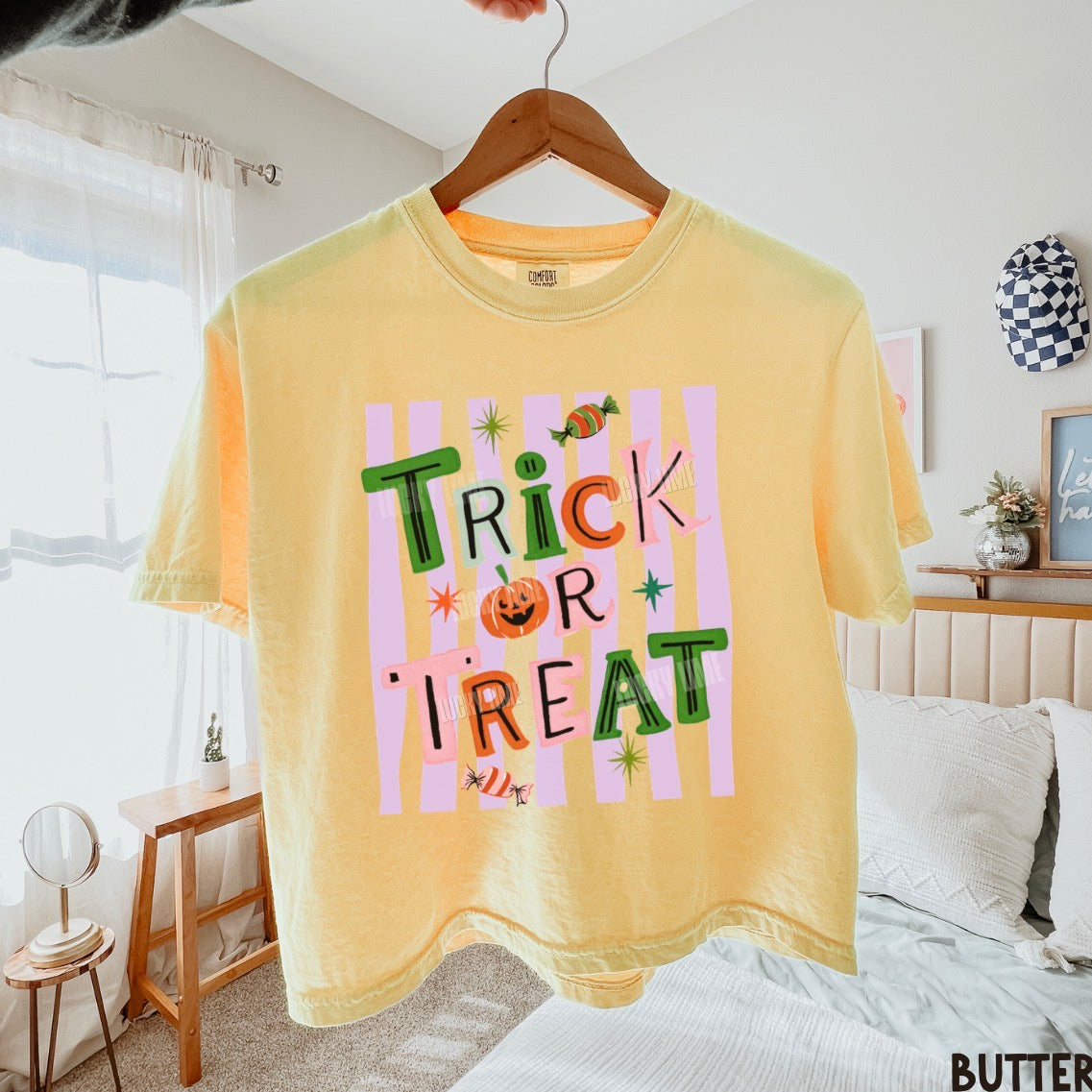 A pale yellow graphic T-shirt hanging on a wooden hanger in a bedroom setting. The shirt features a colorful 'Trick or Treat' design with pink vertical stripes, a jack-o'-lantern, candy, and star accents.