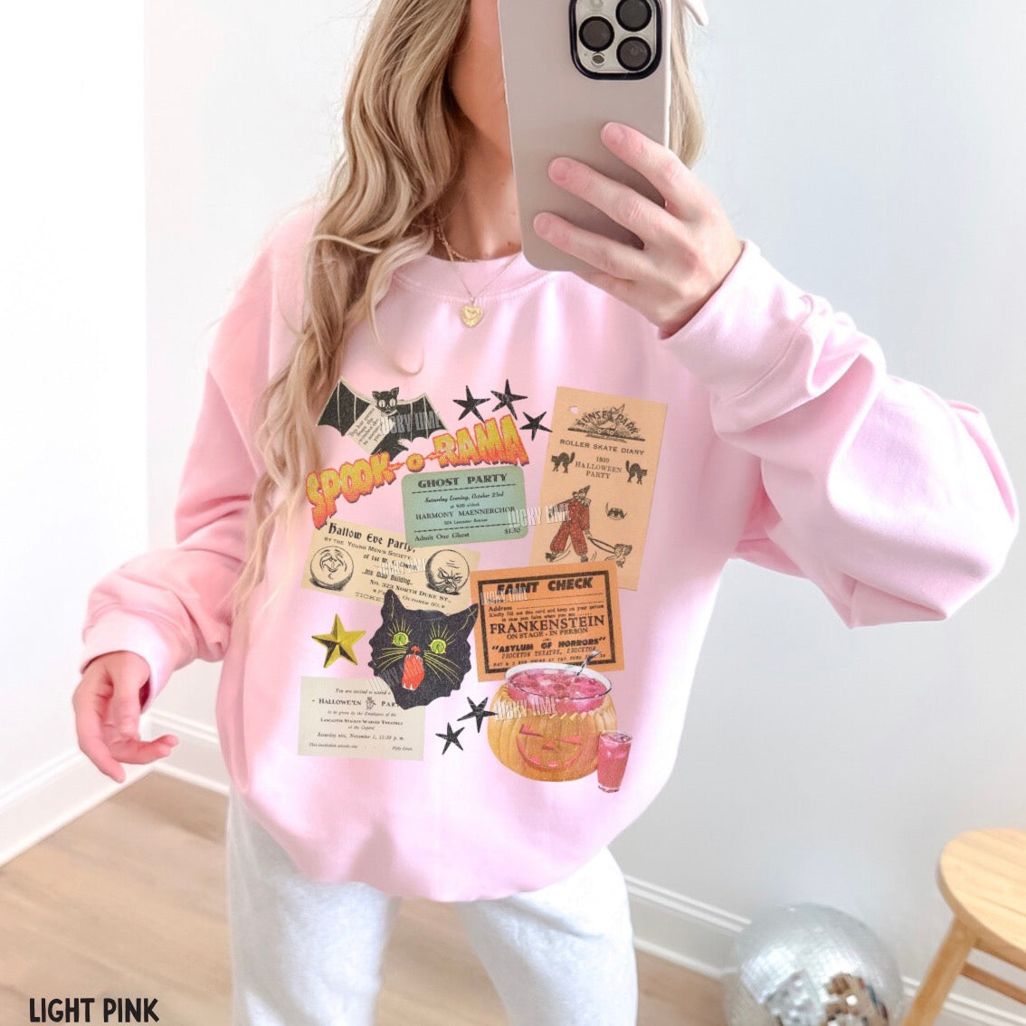 A woman takes a mirror selfie wearing a light pink crewneck sweatshirt with a colorful Halloween-themed graphic print featuring a black cat, candy corn, and party tickets. She is also wearing white sweatpants.