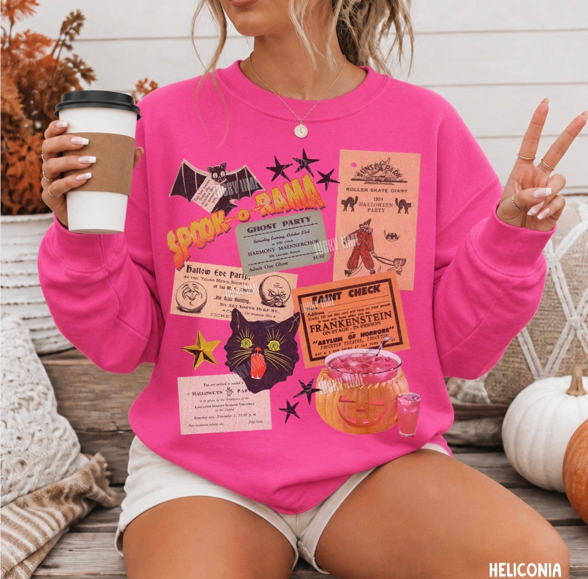 A woman wearing a bright pink crewneck sweatshirt with a colorful Halloween-themed graphic collage, holding a coffee cup and posing with peace signs in an outdoor patio setting.