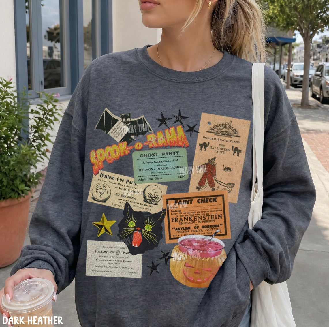 A woman wearing a dark heather gray crewneck sweatshirt with a collage of vintage-style Halloween-themed graphic patches, including 'Spooktacular', 'Haunted Mansion', 'Frankenstein', and a black cat. She is carrying a white shoulder bag and holding a drink, set against an outdoor street background.