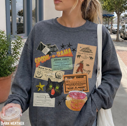 A woman wearing a dark heather gray crewneck sweatshirt with a collage of vintage-style Halloween-themed graphic patches, including 'Spooktacular', 'Haunted Mansion', 'Frankenstein', and a black cat. She is carrying a white shoulder bag and holding a drink, set against an outdoor street background.