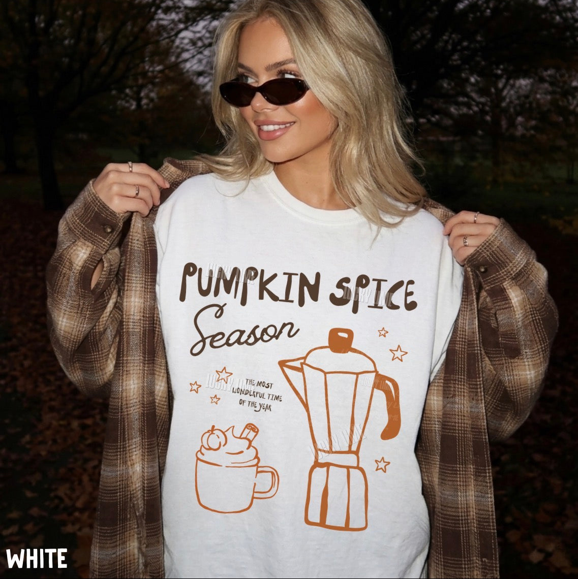 A woman outdoors wearing a white graphic T-shirt with 'Pumpkin Spice Season' text and pumpkin spice latte illustrations, styled with a brown plaid shirt draped off her shoulders and dark sunglasses.