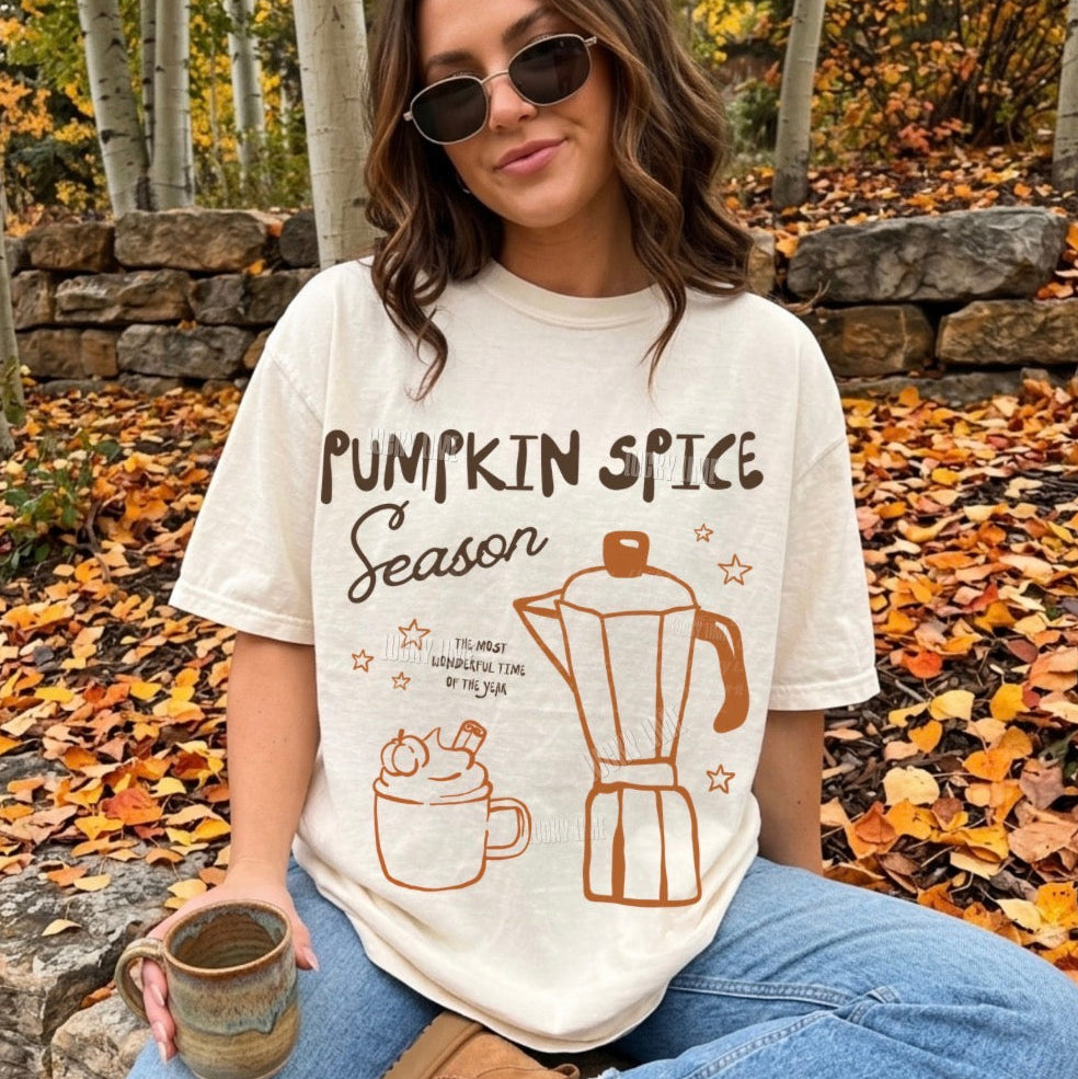 A woman wearing a cream-colored graphic t-shirt with 'Pumpkin Spice Season' and illustrations of a mug and coffee maker, sitting outdoors in a wooded area with fallen leaves.