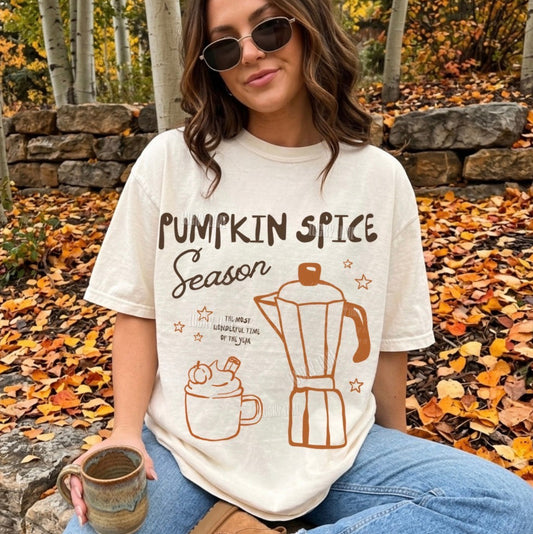 A woman wearing a cream-colored graphic t-shirt with 'Pumpkin Spice Season' and illustrations of a mug and coffee maker, sitting outdoors in a wooded area with fallen leaves.