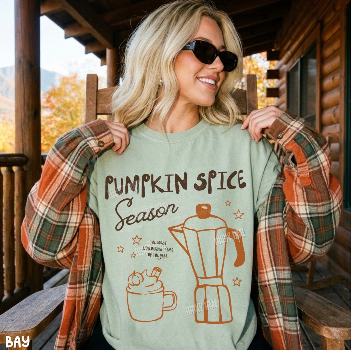A woman wearing a light sage green graphic t-shirt that reads 'PUMPKIN SPICE Season' with a coffee mug and moka pot illustration, paired with an open rust-orange plaid flannel shirt, sitting on a wooden porch chair.