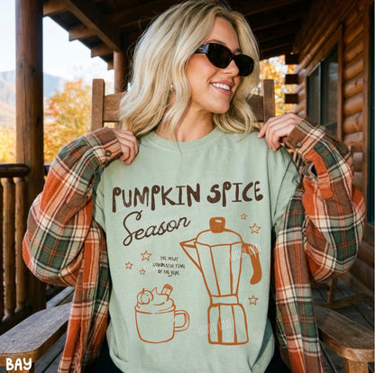 A woman wearing a light sage green graphic t-shirt that reads 'PUMPKIN SPICE Season' with a coffee mug and moka pot illustration, paired with an open rust-orange plaid flannel shirt, sitting on a wooden porch chair.