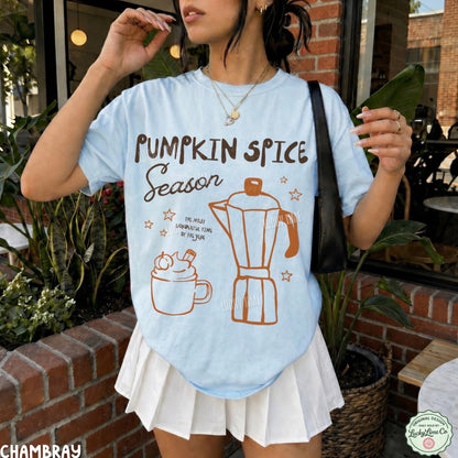 A woman wearing a light blue graphic T-shirt with 'Pumpkin Spice Season' text and illustrations of a coffee mug and moka pot, paired with a white pleated skirt in an outdoor patio setting.