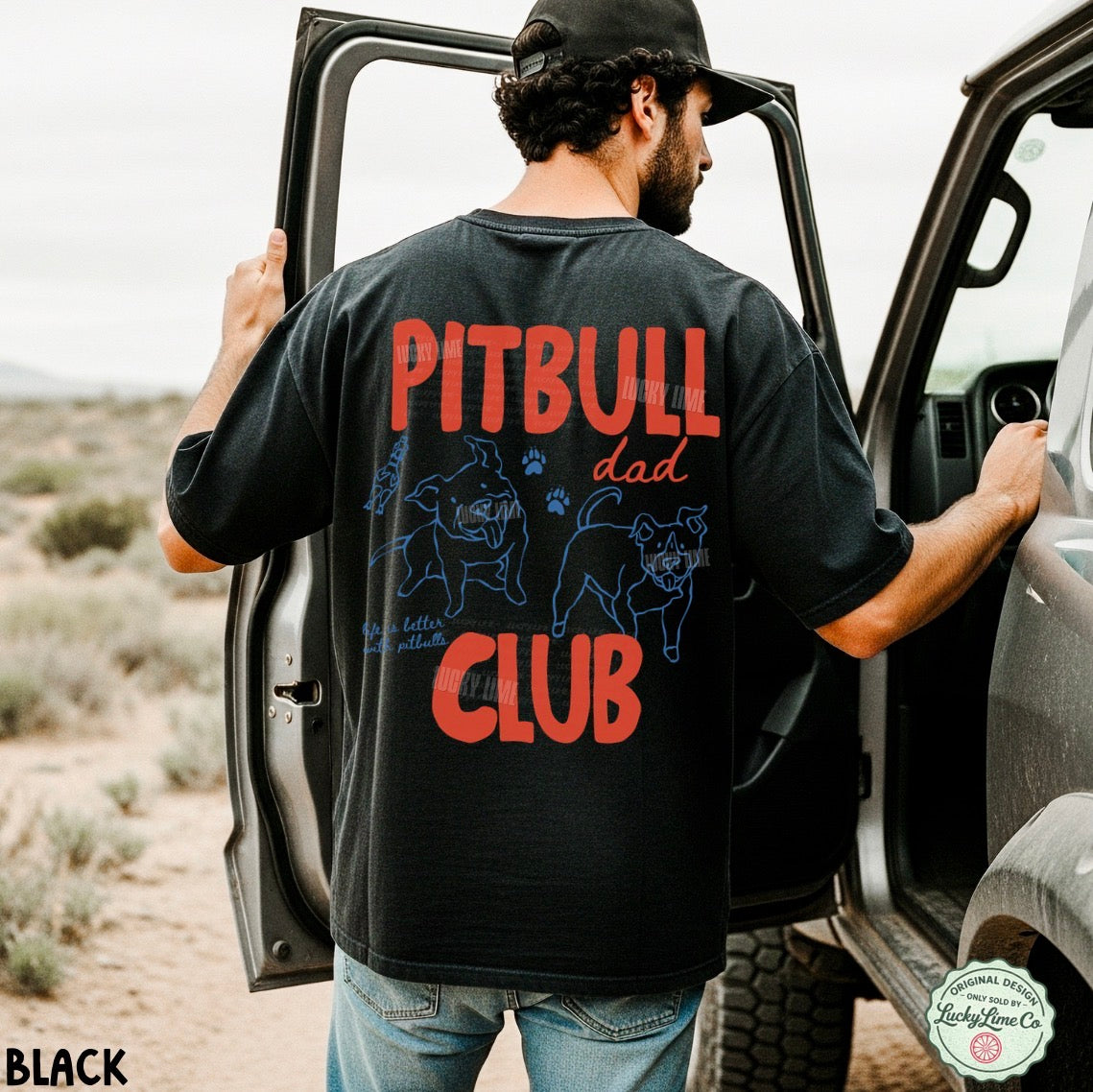 A man wearing a black t-shirt with a red and blue graphic on the back, standing next to an open car door in an outdoor desert setting. The graphic reads 'PITBULL dad CLUB' with illustrations of two pitbull dogs.