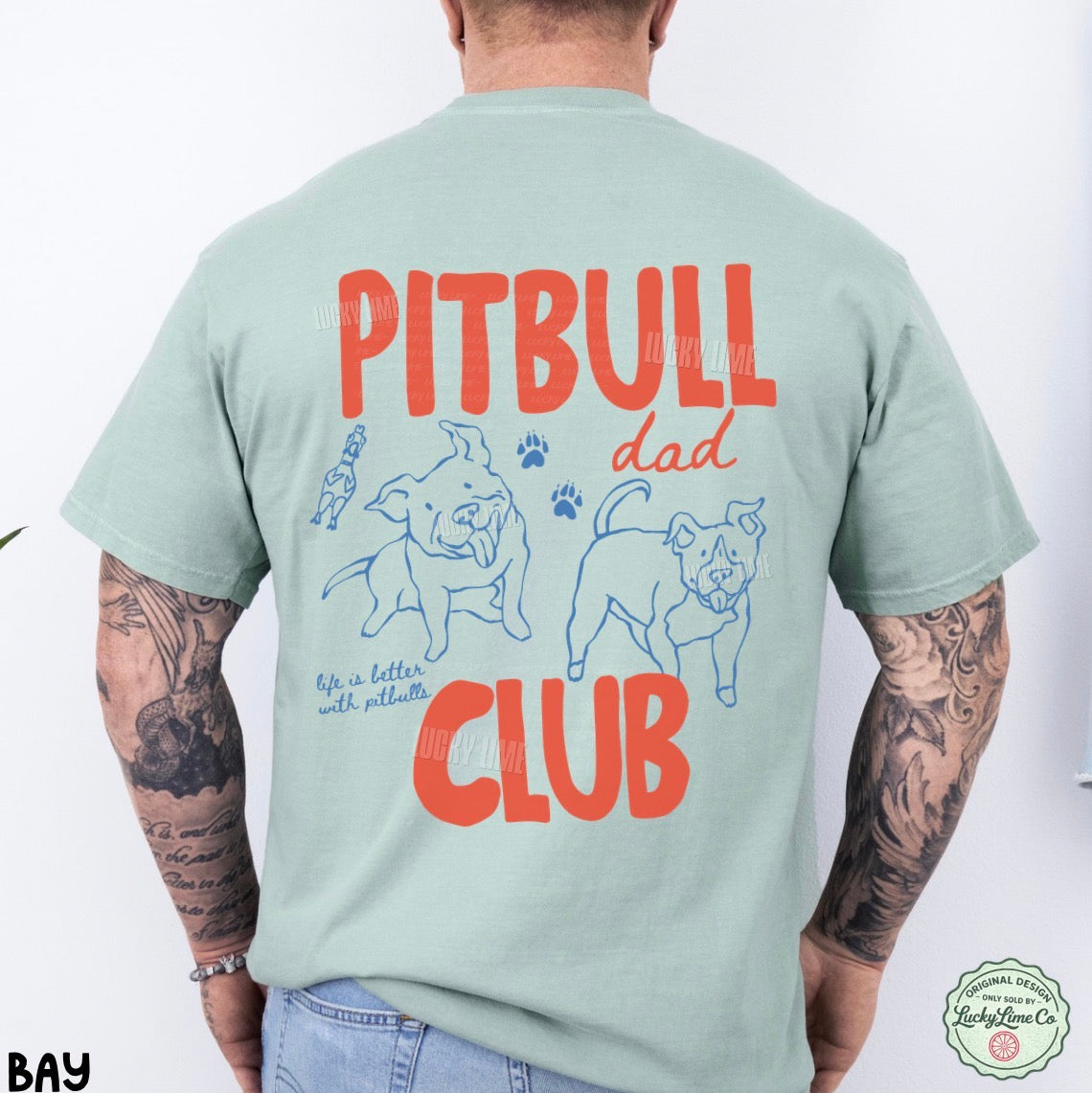 A man viewed from the back wearing a light sage green t-shirt with a large graphic print on the back. The graphic features the words 'PITBULL dad CLUB' in bold red-orange lettering, accompanied by blue line drawings of two pitbull dogs, paw prints, and the phrase 'life is better with pitbulls'.