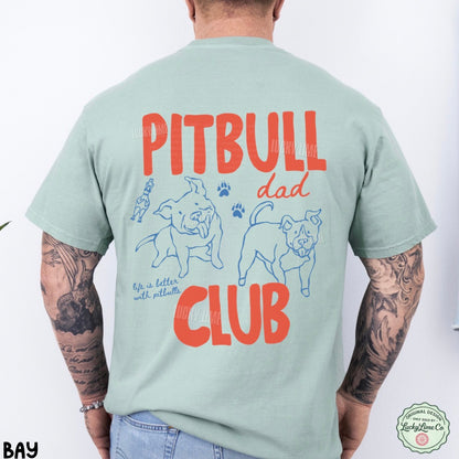 A man viewed from the back wearing a light sage green t-shirt with a large graphic print on the back. The graphic features the words 'PITBULL dad CLUB' in bold red-orange lettering, accompanied by blue line drawings of two pitbull dogs, paw prints, and the phrase 'life is better with pitbulls'.