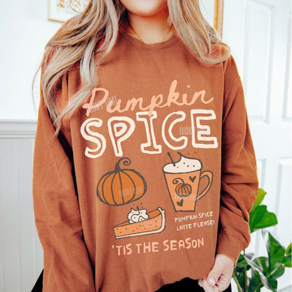 A woman wearing a brown crewneck sweatshirt with a 'Pumpkin Spice' graphic print featuring a pumpkin, latte, and pie, with the text 'Pumpkin Spice Latte Please! Tis the Season'.