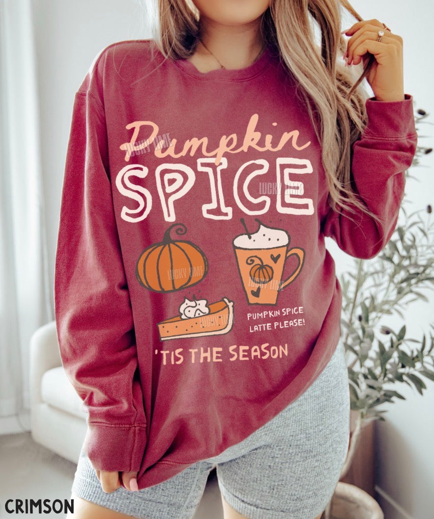 A woman wearing a long-sleeve crewneck sweatshirt in a deep crimson color, featuring a 'Pumpkin Spice' graphic with illustrations of a pumpkin, pie, and a mug. The image is cropped from the chest down, showing gray heather shorts, with a soft-focus indoor background.