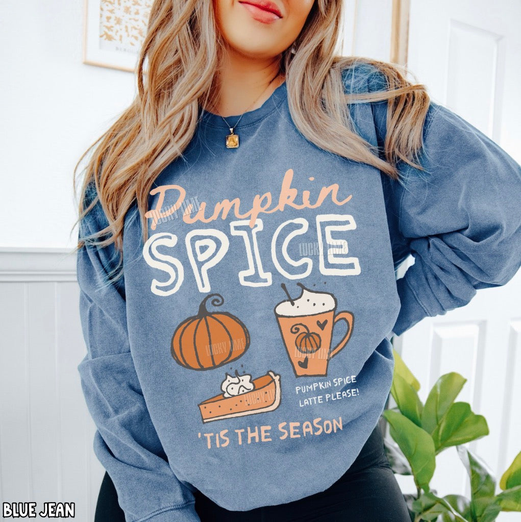 A woman wearing a heathered blue sweatshirt with a 'Pumpkin Spice' graphic print featuring a pumpkin, latte, and pie, in a bright indoor setting.