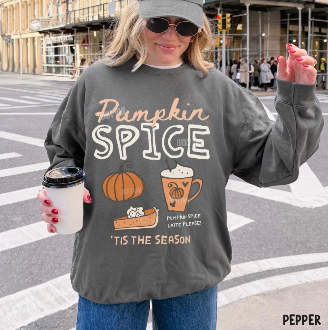 A woman wearing a dark gray graphic sweatshirt that reads 'Pumpkin Spice' with illustrations of a pumpkin, a coffee cup, and a slice of pie, standing on a city street crosswalk.