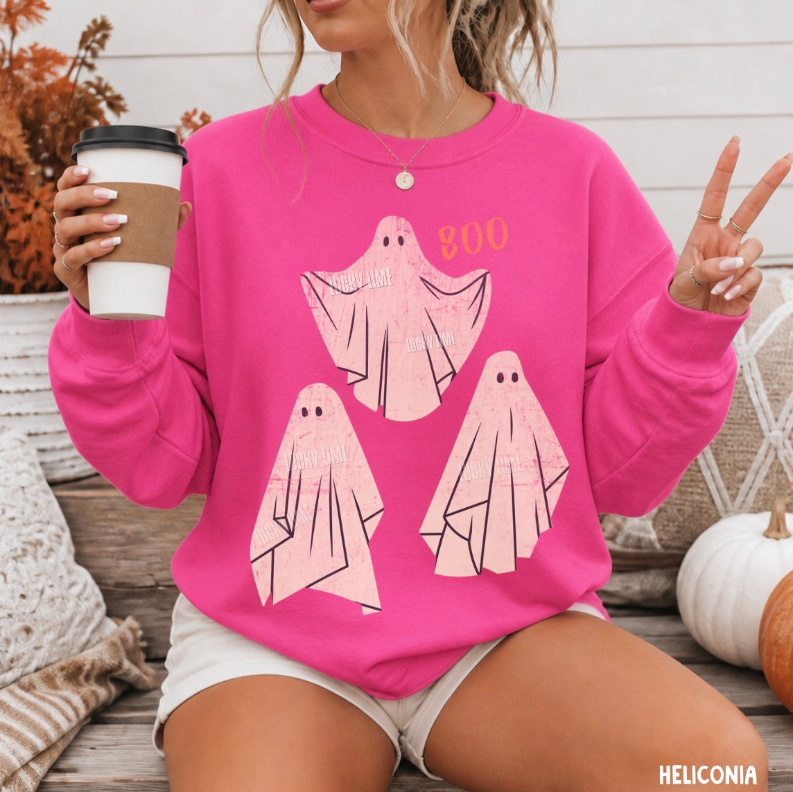 A woman wearing a bright pink crewneck sweatshirt with a graphic of three light pink ghosts and the word 'BOO' in orange, holding a coffee cup and posing with peace signs.