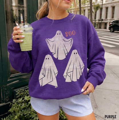 A woman wearing a purple crewneck sweatshirt with a graphic of three white ghosts and the word 'BOO' in orange, holding a green iced drink on a city sidewalk.