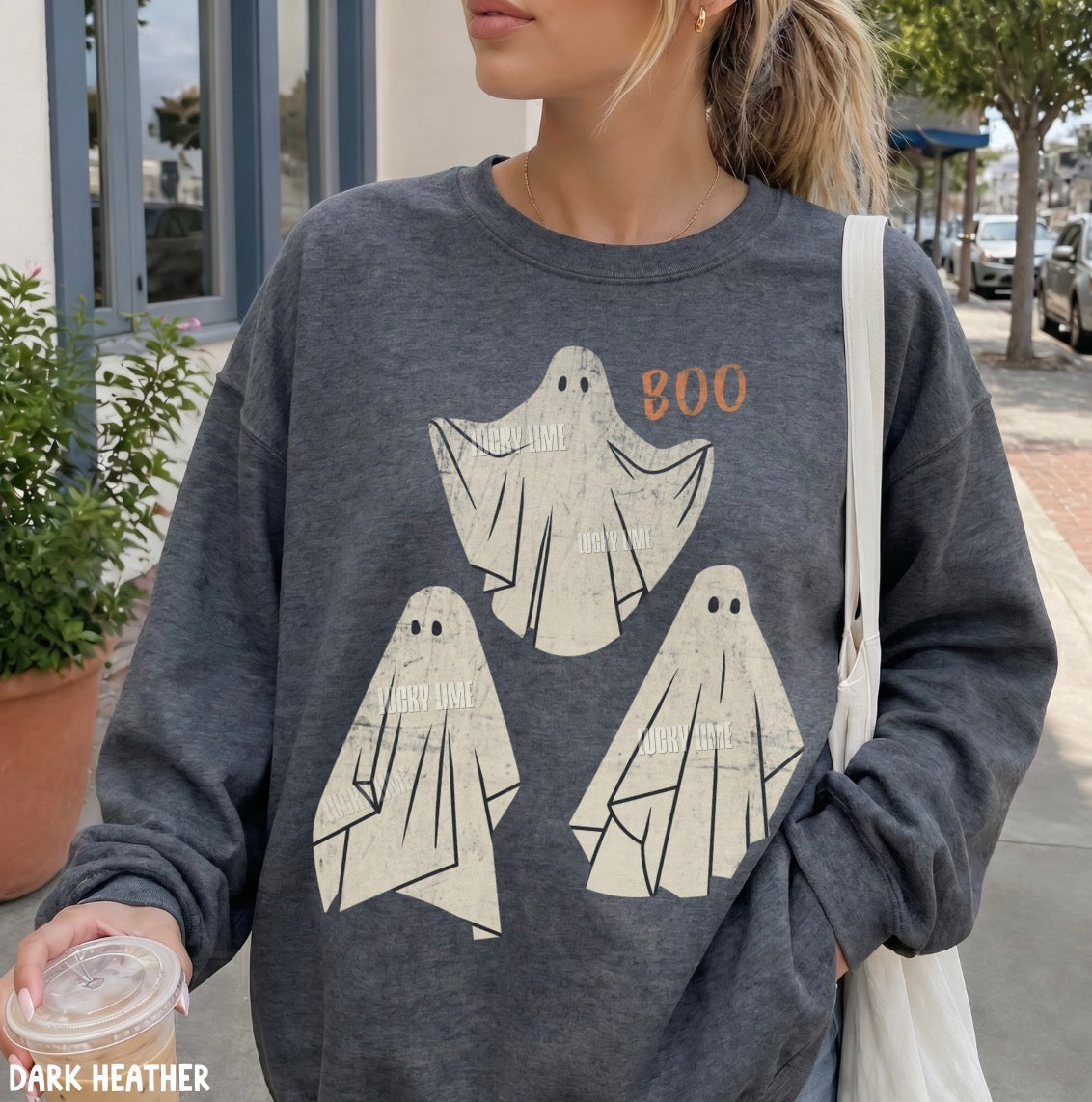 A woman wearing a dark heather gray crewneck sweatshirt with a graphic of three cream-colored ghosts and the word 'BOO' in orange. She is holding a takeaway coffee cup on a sunny outdoor sidewalk.