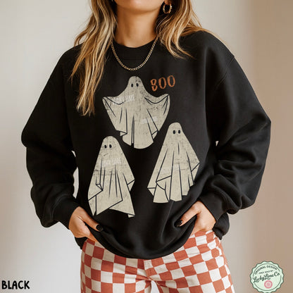 A woman wearing a black crewneck sweatshirt with a distressed graphic of three white ghosts and the word 'BOO' in orange. She is also wearing red and white checkered pants, standing against a neutral beige background.