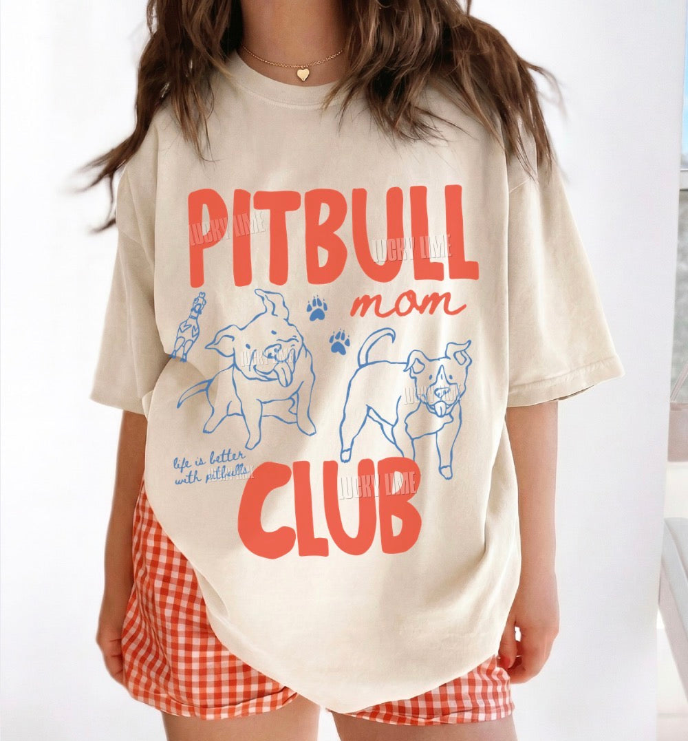 A woman wearing an oversized cream-colored t-shirt with a red and blue graphic that reads 'PITBULL mom CLUB' with cartoon dog illustrations. She is also wearing red and white gingham shorts.