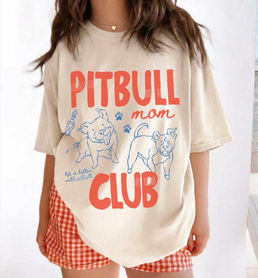 A woman wearing an oversized cream-colored t-shirt with a red and blue graphic that reads 'PITBULL mom CLUB' with cartoon dog illustrations. She is also wearing red and white gingham shorts.