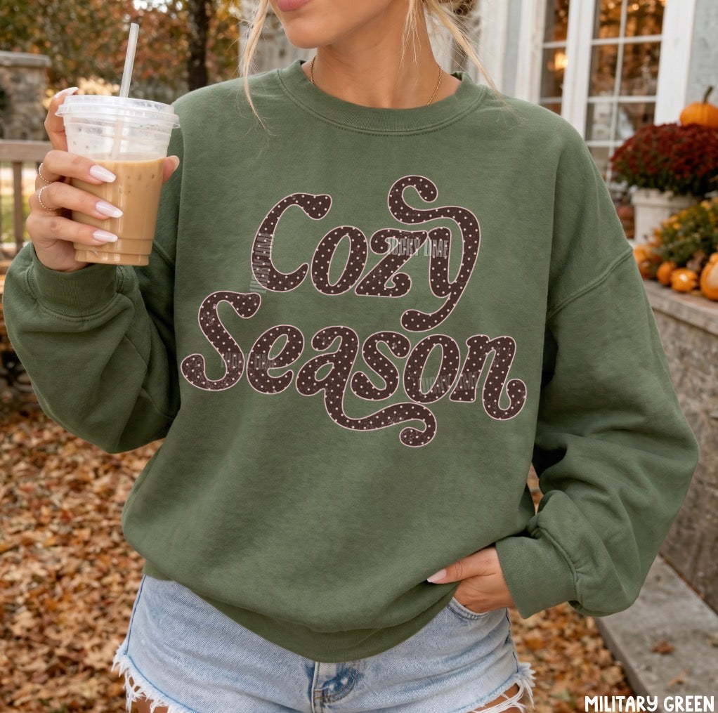 A woman wearing an olive green sweatshirt with 'Cozy Season' written across the front in a glittery dark brown script. She is holding an iced coffee outdoors in front of a house during autumn, with fallen leaves on the ground and pumpkins in the background.