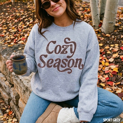 A woman wearing a light grey sweatshirt with 'Cozy Season' printed in brown glitter-style lettering, sitting outdoors on a stone wall surrounded by fallen autumn leaves.