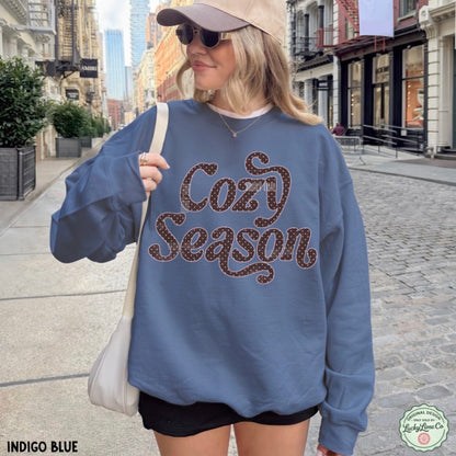 A woman wearing an oversized indigo blue sweatshirt with 'Cozy Season' written across the front in brown glitter-style lettering, standing on a city street. She is also wearing a beige cap, sunglasses, black shorts, and carrying a light beige shoulder bag.