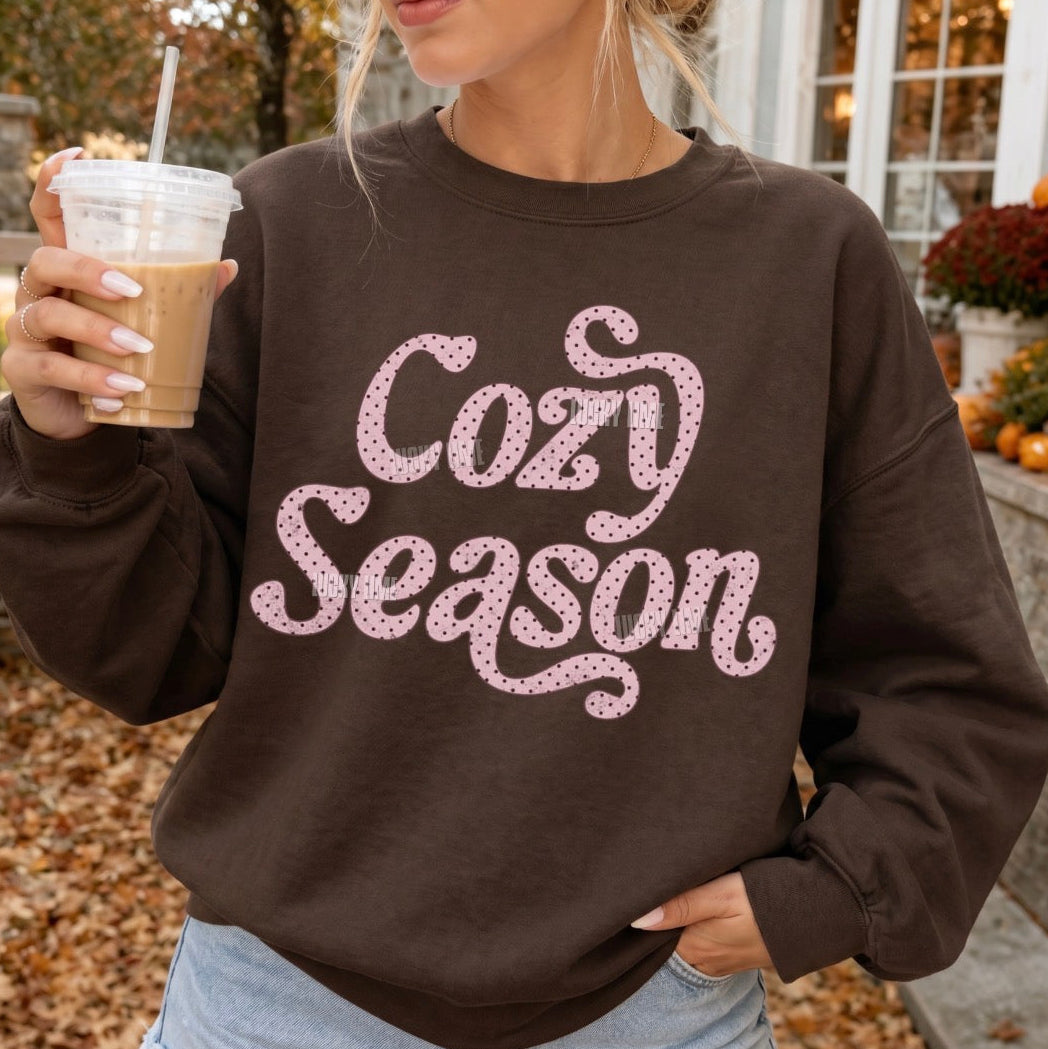 A woman wearing a dark brown sweatshirt with 'Cozy Season' printed in pink glitter-style lettering, holding an iced coffee outdoors in front of a house.