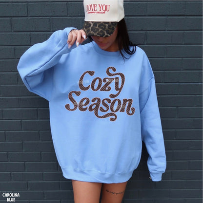A person wearing an oversized light blue sweatshirt with 'Cozy Season' in brown leopard-print lettering, standing against a dark gray brick wall. The person is also wearing a beige cap with leopard print and red text.
